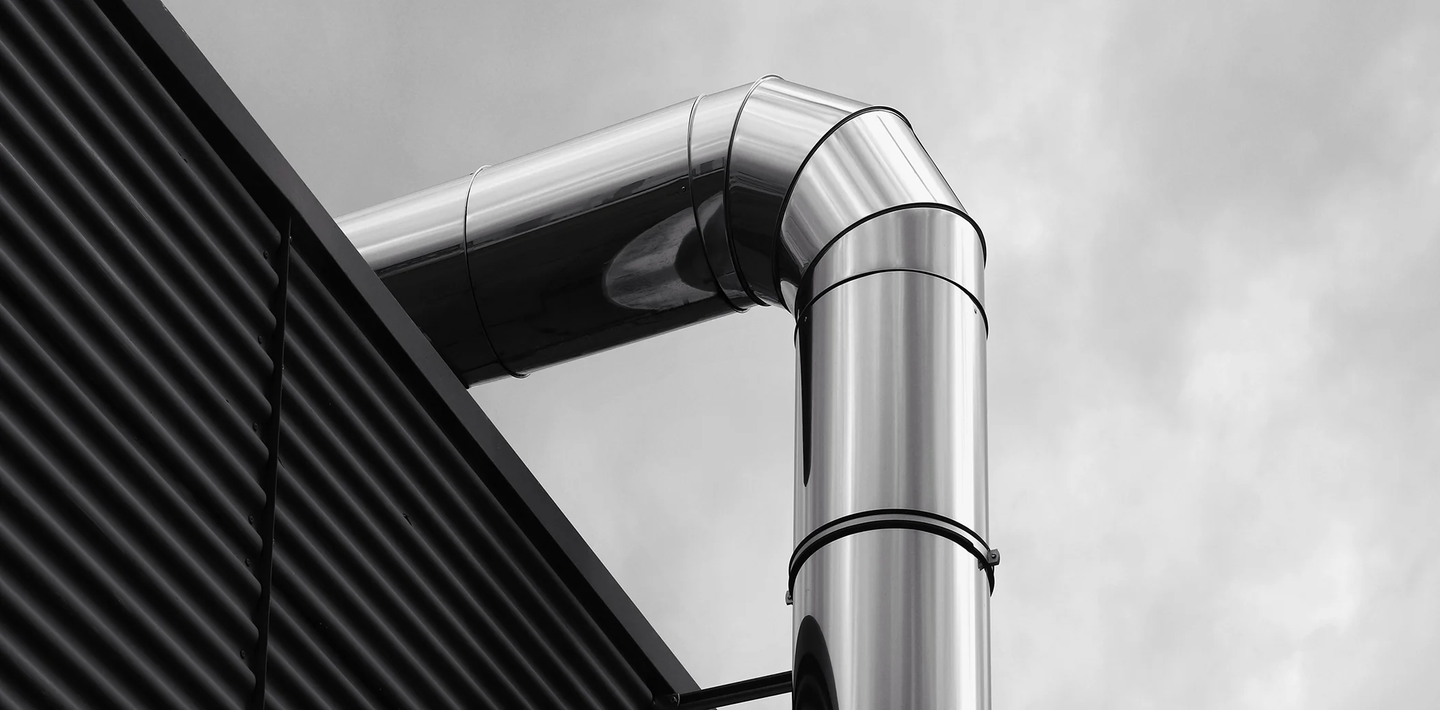 Szczegółowy widok błyszczącego kolana rury w czarno-białych barwach. Segmentowe kolano rury o wysokim połysku na tle nieba i fasady budynku. Rury są nitowane i mają widoczne przetłoczenia. Kontrastowe oświetlenie podkreśla teksturę.
